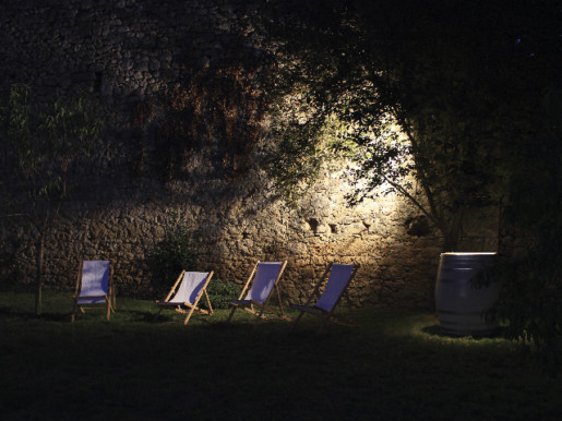 Dans ce jardin de Gironde, une barrique sert de mange-debout. Un luminaire révèle un mur en pierre, ordinaire de jour : l'ombre portée du végétal accentue sa présence.